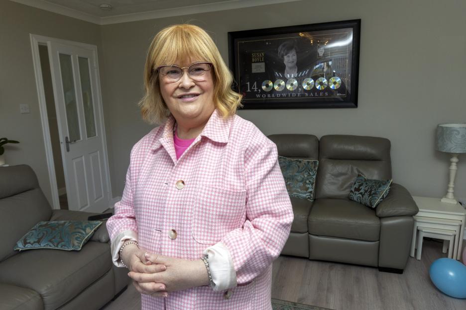Susan Boyle posing in her new home, with a framed display of her album sales on the wall.