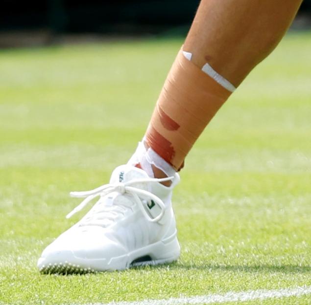 Close-up of a tennis player's leg with an ankle taped with medical tape, wearing a white tennis shoe, on a green grass court.