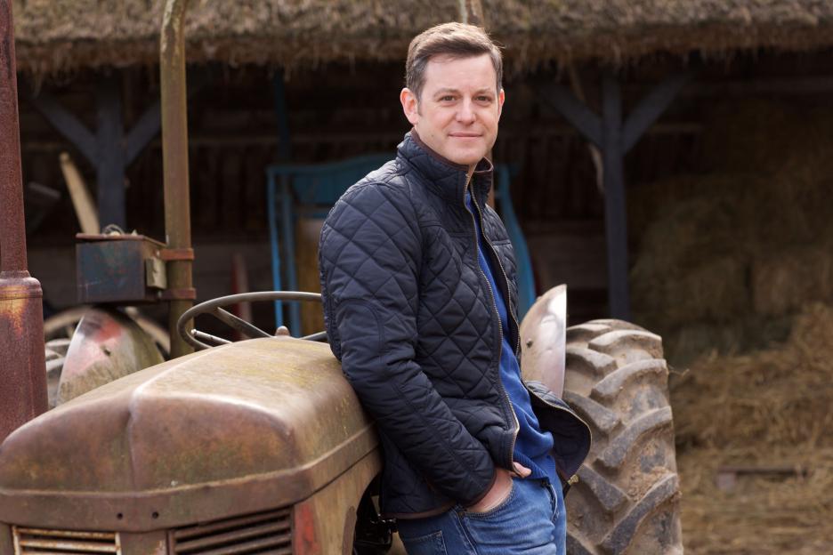 Matt Baker leaning on a rusty tractor in front of a thatched building.
