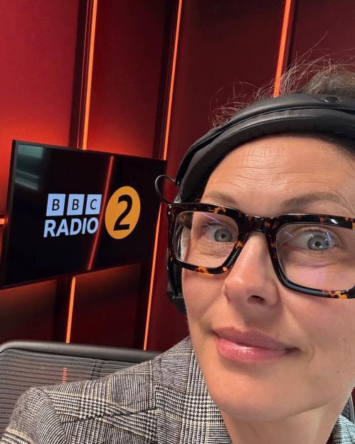 Emma Willis, wearing large tortoise-shell glasses and headphones, taking a selfie in front of a monitor displaying the BBC Radio 2 logo.