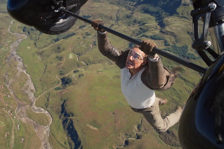 Tom Cruise hanging from a plane with green hills and a river below.