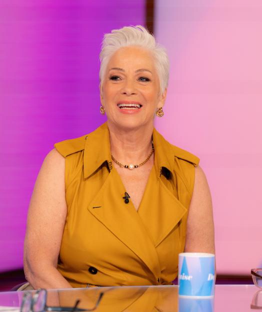 Denise Welch, a woman with short white hair, smiling and wearing a mustard yellow top, gold necklace, and earrings.
