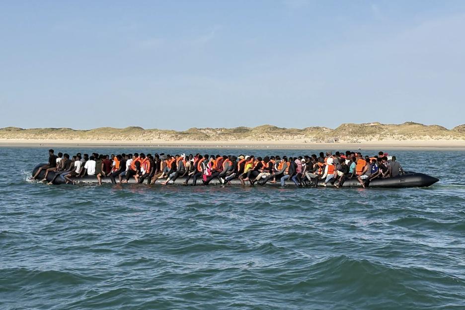 Migrants in an overcrowded dinghy boat crossing the English Channel, with sandy dunes in the background.