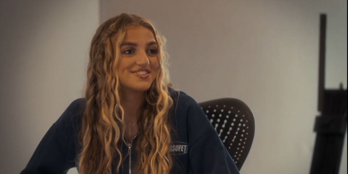 A young woman with long, curly blonde hair smiles and looks to the right.