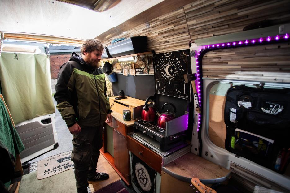 A man stands inside his converted minibus, looking at two red kettles on a portable stove.