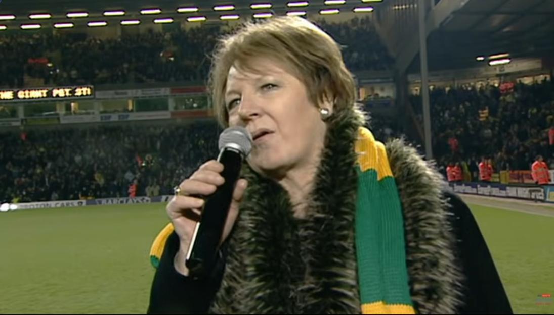 Delia Smith speaks into a microphone on a football pitch, wearing a fur boa and a yellow and green scarf, with fans in the stands behind her.