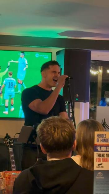 Jamie Jewitt performing a song with a microphone in front of a screen displaying a soccer game.