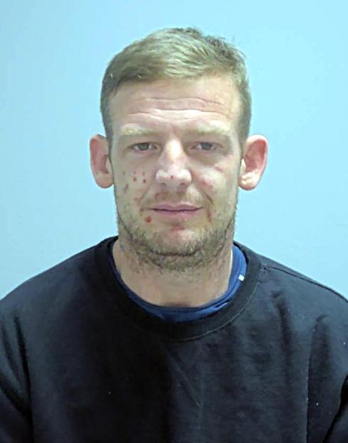 Mugshot of John Scott, a man with light stubble and acne scars on his face, wearing a black long-sleeved shirt.