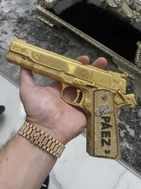 Hand holding an ornate gold pistol with "PÁEZ" and a cowboy hat emblem encrusted in diamonds.