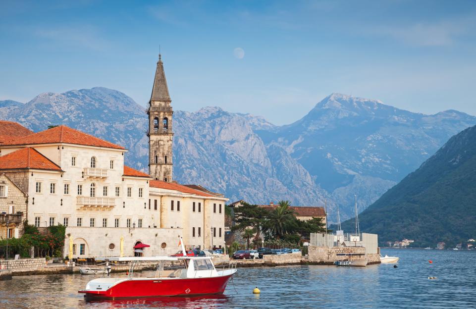 Old Perast town in Kotor Bay, Montenegro, featuring historic buildings with red roofs, a prominent bell tower, and a red and white boat in the foreground, all set against a backdrop of mountains and a moon in the sky.