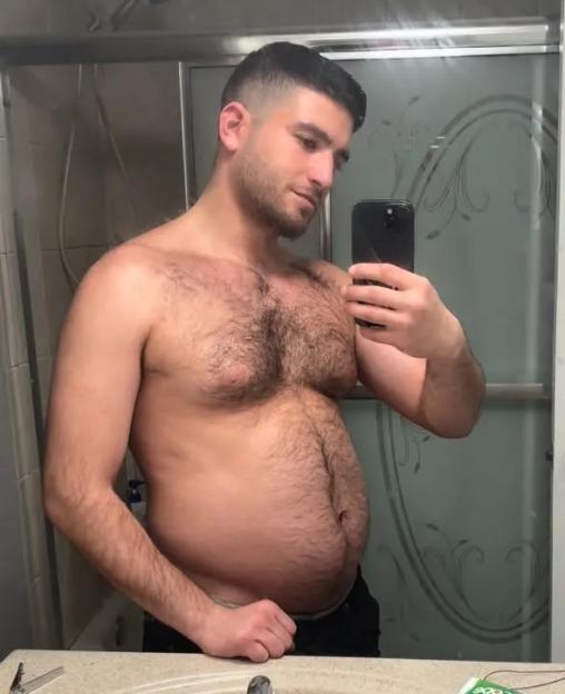Man with a beard taking a selfie in the bathroom mirror, showing his torso and stomach.
