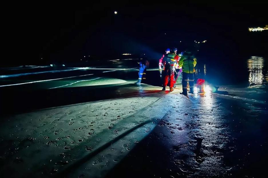 Helicopter spotlight searching over dark water near a lit coastline at night.