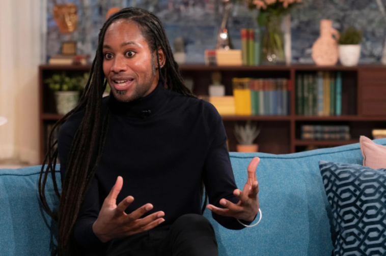 A person with dreadlocks and a black turtleneck sits on a blue couch, gesturing with their hands.