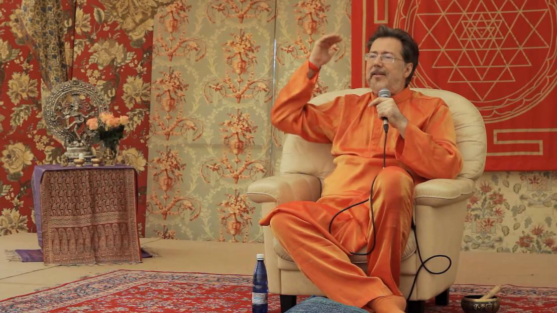 Narcis Tarcau, wearing an orange tunic, seated in a cream chair speaking into a microphone.