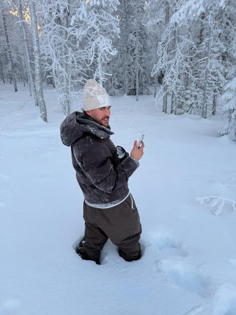 A man in a winter hat and coat kneels in deep snow, holding a smartphone, with snow-covered trees in the background.