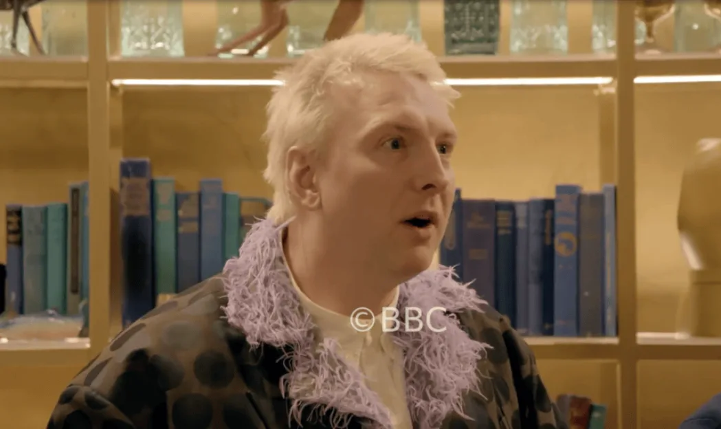 Joe Lycett in The Traitors, wearing a patterned jacket with a purple feather trim.