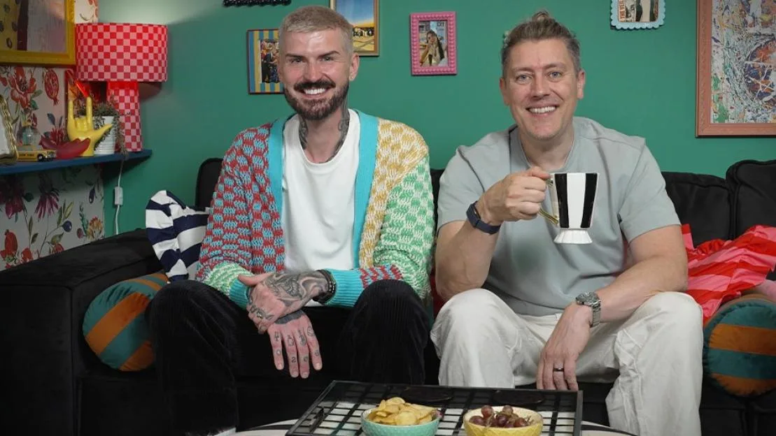 Two men on a couch, one in a colorful cardigan and the other holding a striped mug.