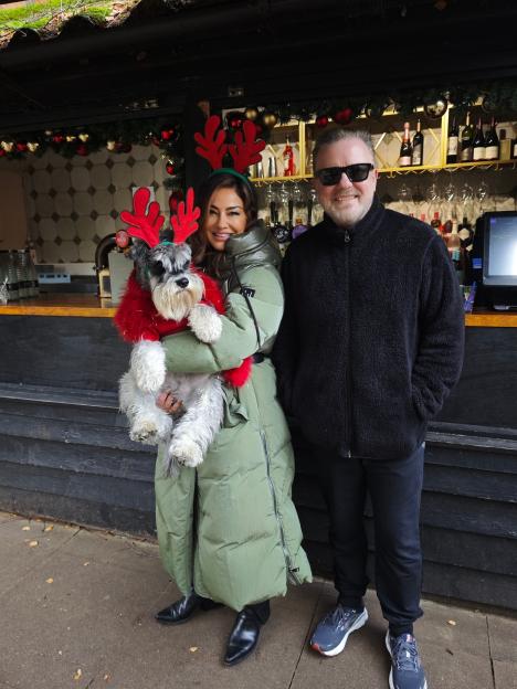 Ricky Gervais and a woman holding a dog wearing a red Christmas outfit.