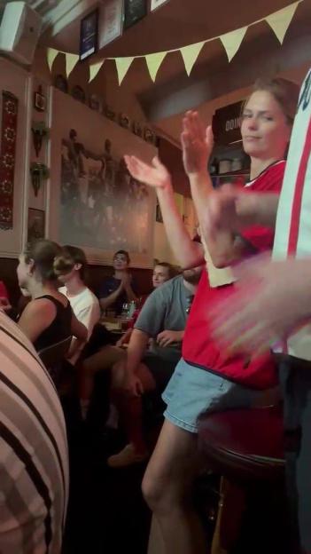 A crowd of people in a dimly lit room, with one woman in a red shirt clapping, and banners hanging overhead.