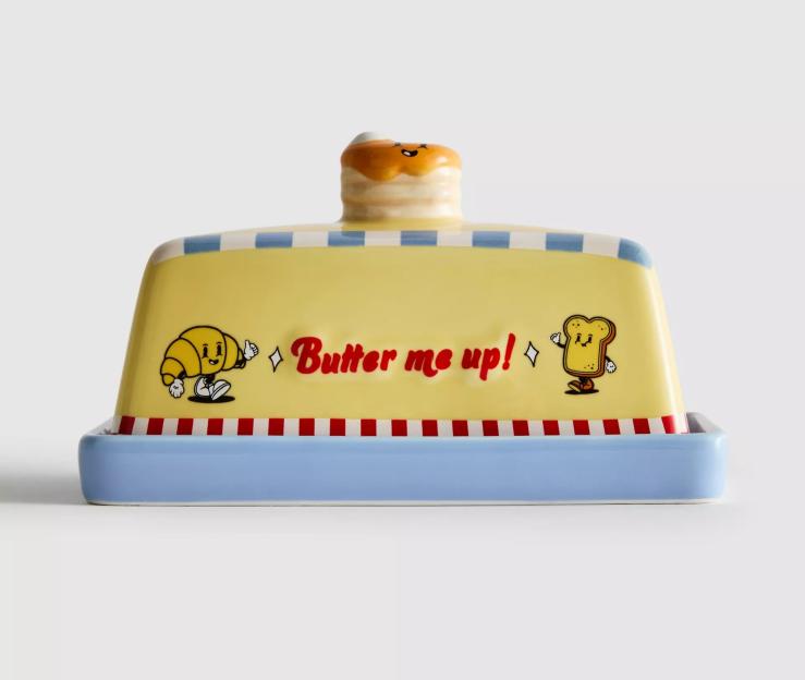 A yellow ceramic butter dish with blue and red stripes, featuring cartoon croissant and toast characters, and the text "Butter me up!".