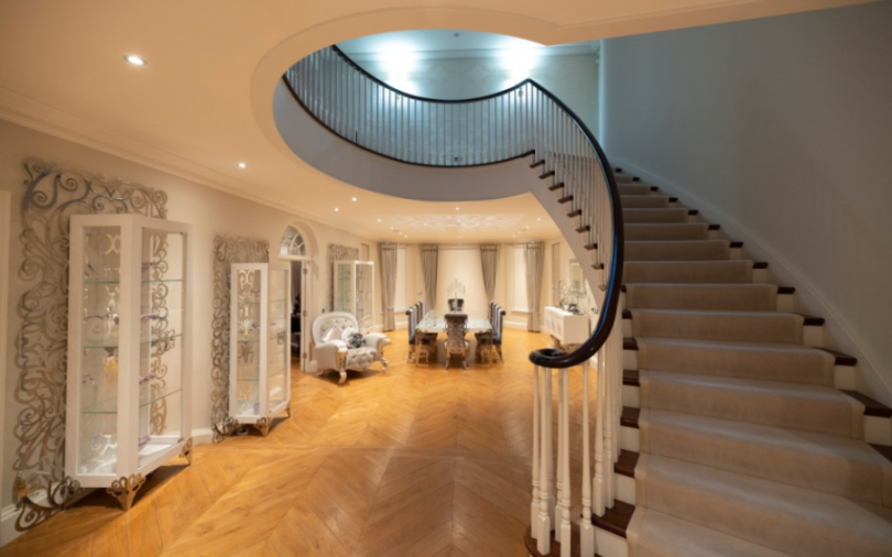 A luxurious foyer with a grand curved staircase leading upstairs, a dining room with a long table and chairs in the background, and decorative glass display cabinets on the left.