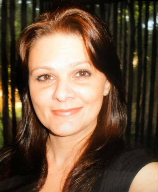 A woman with long dark brown hair and brown eyes smiles faintly, wearing a dark top, with a blurred background of vertical dark stripes.