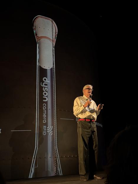 Man presenting on stage next to a tall screen displaying "Dyson camera RFID".