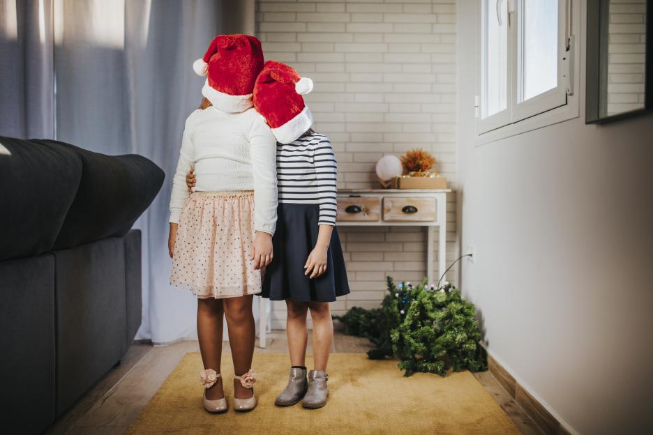 Two girls wearing Santa hats that obscure their faces, one with her arm around the other, in a room with a fallen Christmas tree.