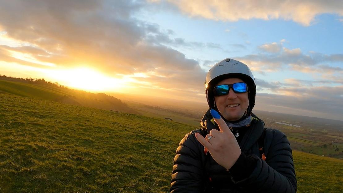 Man in helmet and sunglasses making a hand gesture with a bright sunset behind him over a green hillside.