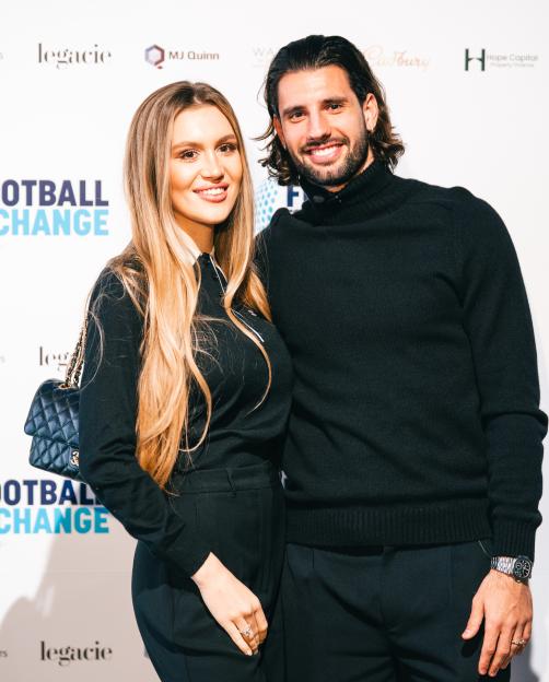 A man and a woman in black standing in front of a white wall with the logos "legacie", "MJ Quinn", "Cadbury", "Hope Capital Property Finance", and "Football for Change" printed on it.