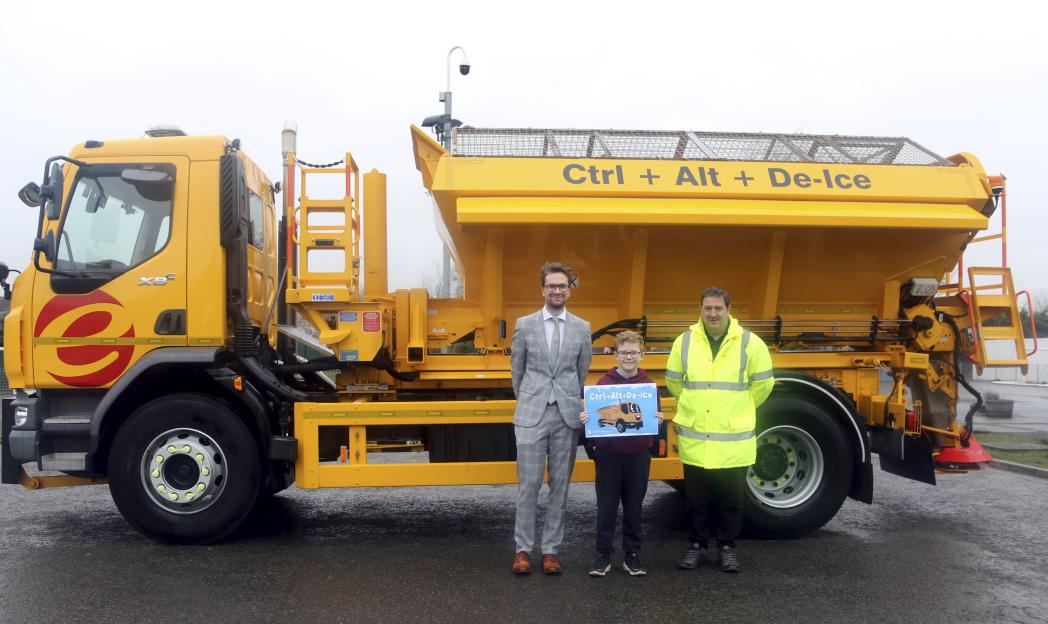 A primary student, Daniel, holding a sign with a cartoon gritter, stands next to a man in a suit and a gritter driver, in front of a gritter truck named "Ctrl + Alt + De-Ice".