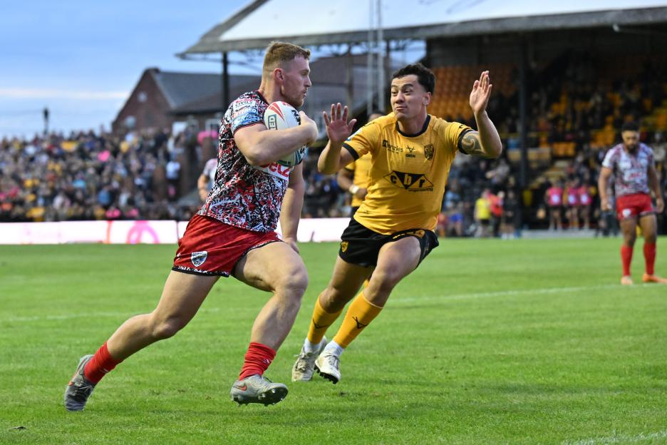 A Rugby player in a red and white patterned jersey runs with the ball while a player in a yellow jersey defends.