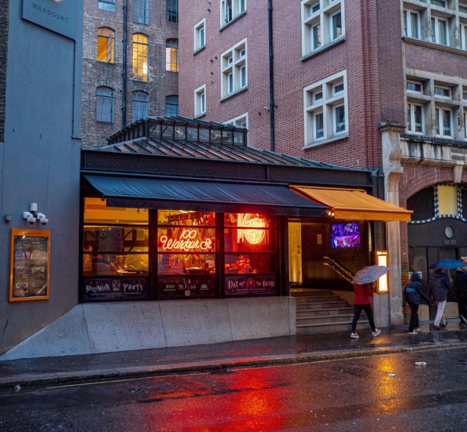 The exterior of 100 Wardour St Restaurant and Bar in London's Soho district, featuring a neon "100 Wardour St" sign in its window and reflections on the wet street.