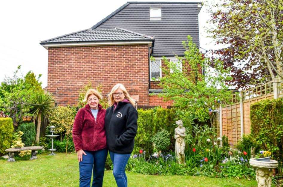 Two women standing in a garden with a house in the background featuring a large dormer on its roof.