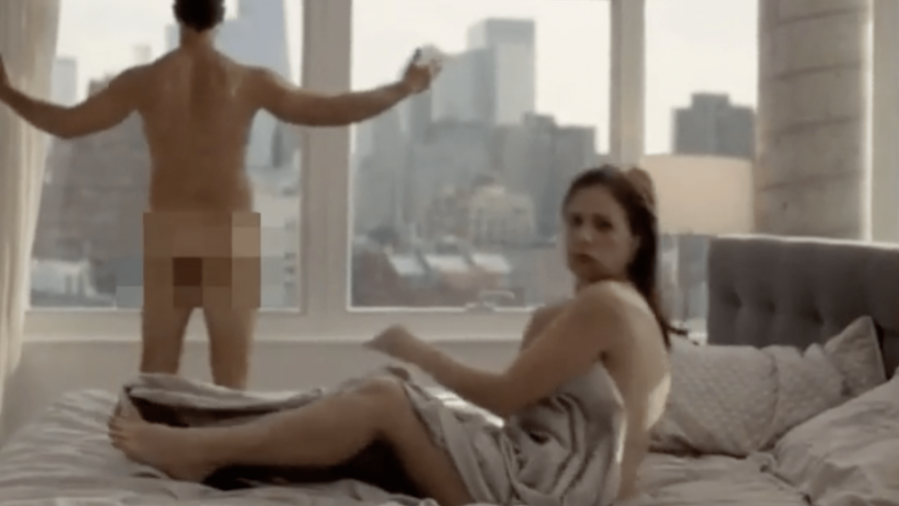 Nude man standing at a window while a woman is in bed with a blanket.