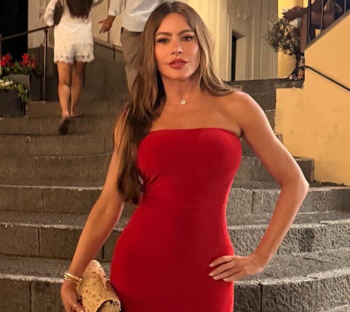 Sofia Vergara in a strapless red dress, holding a gold clutch, standing on stone steps
