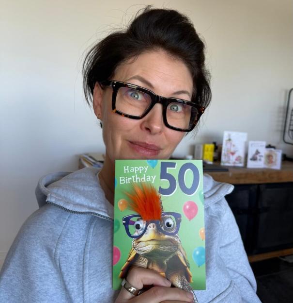 Emma Willis holds up a birthday card that reads "Happy Birthday 50" with a picture of a turtle wearing glasses and an orange mohawk.