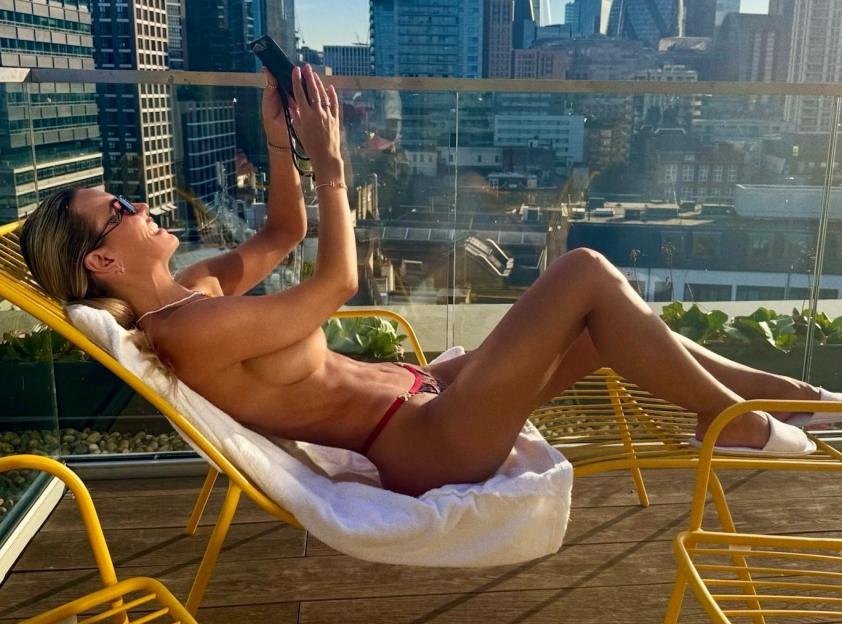 Christine McGuinness relaxing topless on a deck chair, holding a phone, with a city skyline in the background.
