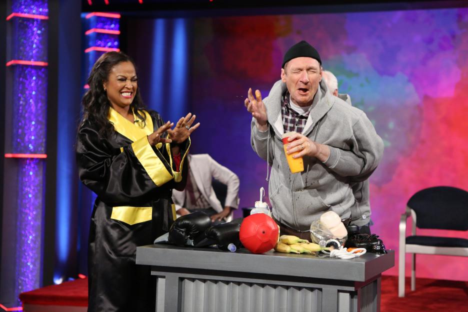 Laila Ali in a black and yellow robe and Ryan Stiles with orange liquid on his face on the show Whose Line is it Anyway?