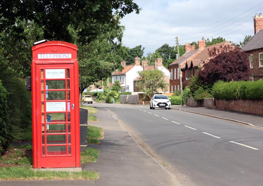 A red telephone booth on the side of a road in a village, with houses and trees in the background.