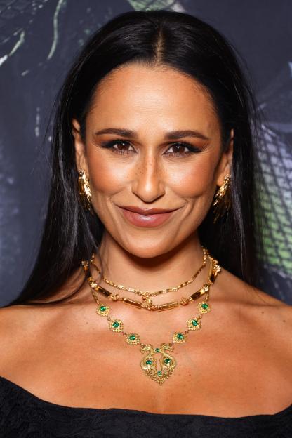 Close-up of a woman with dark hair, a tan complexion, and freckles, smiling at the camera while wearing gold earrings and a gold necklace with green gems.