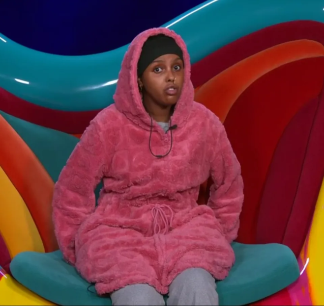 A young woman in a pink fluffy hooded robe, black beanie, and gray pants looks directly at the camera with a surprised expression.