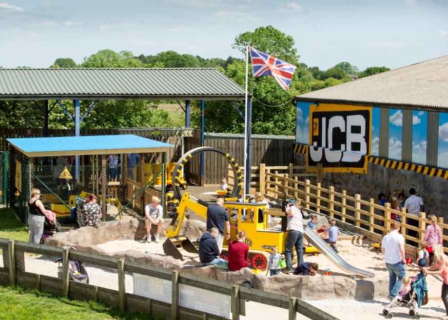 Children and adults play in a sandbox playground with yellow digger toys and a climbing wall, under a Union Jack flag.
