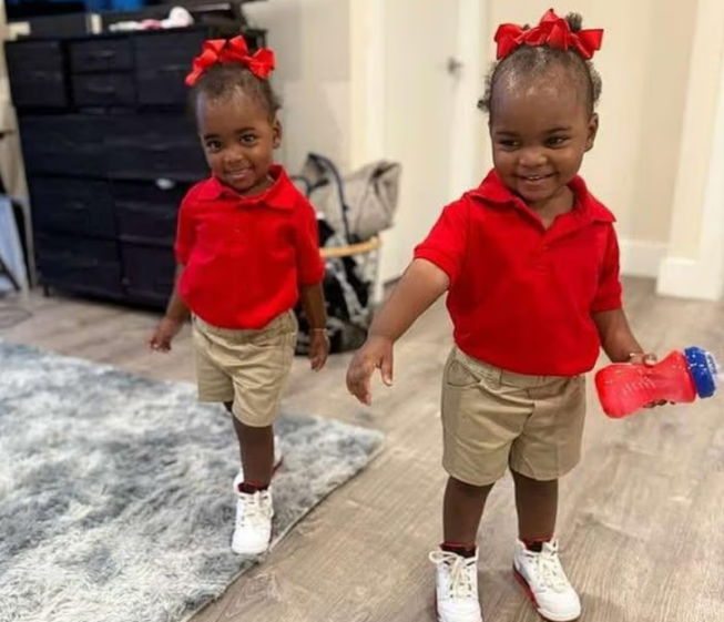 Two toddlers wearing matching red shirts and khaki shorts.
