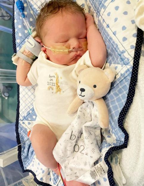 A baby with a nasal feeding tube and arm restraint, wearing a onesie that says "born in 2022," lies next to a teddy bear on a blue and white heart-patterned blanket.