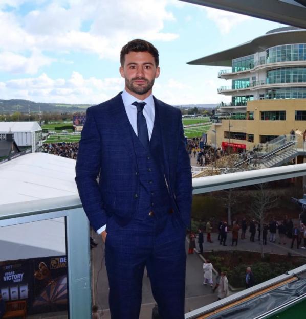 A man in a blue plaid three-piece suit stands on a balcony overlooking a racecourse and a building.
