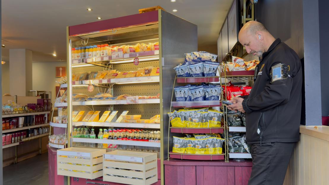 A security guard looking at his phone next to food and drink shelves in a Costa Coffee shop.