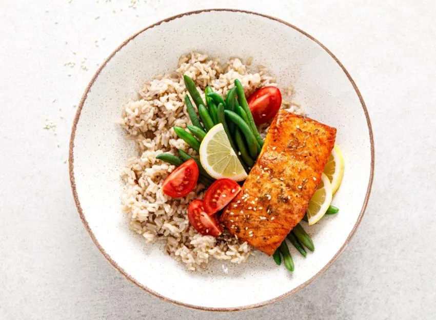 Top-down view of a bowl containing a baked salmon fillet, rice, green beans, cherry tomatoes, and lemon slices.