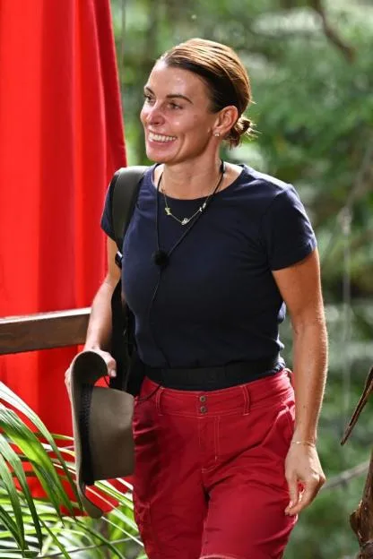 Coleen Rooney smiling while wearing a dark blue t-shirt and red pants.