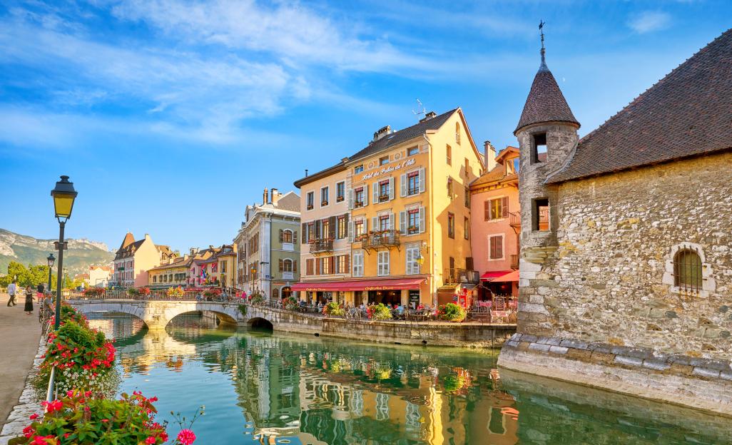 The Palais de l'Isle and Canal de Thiou in Annecy, France, featuring boats, bridges, historic buildings, and floral displays along the canal.